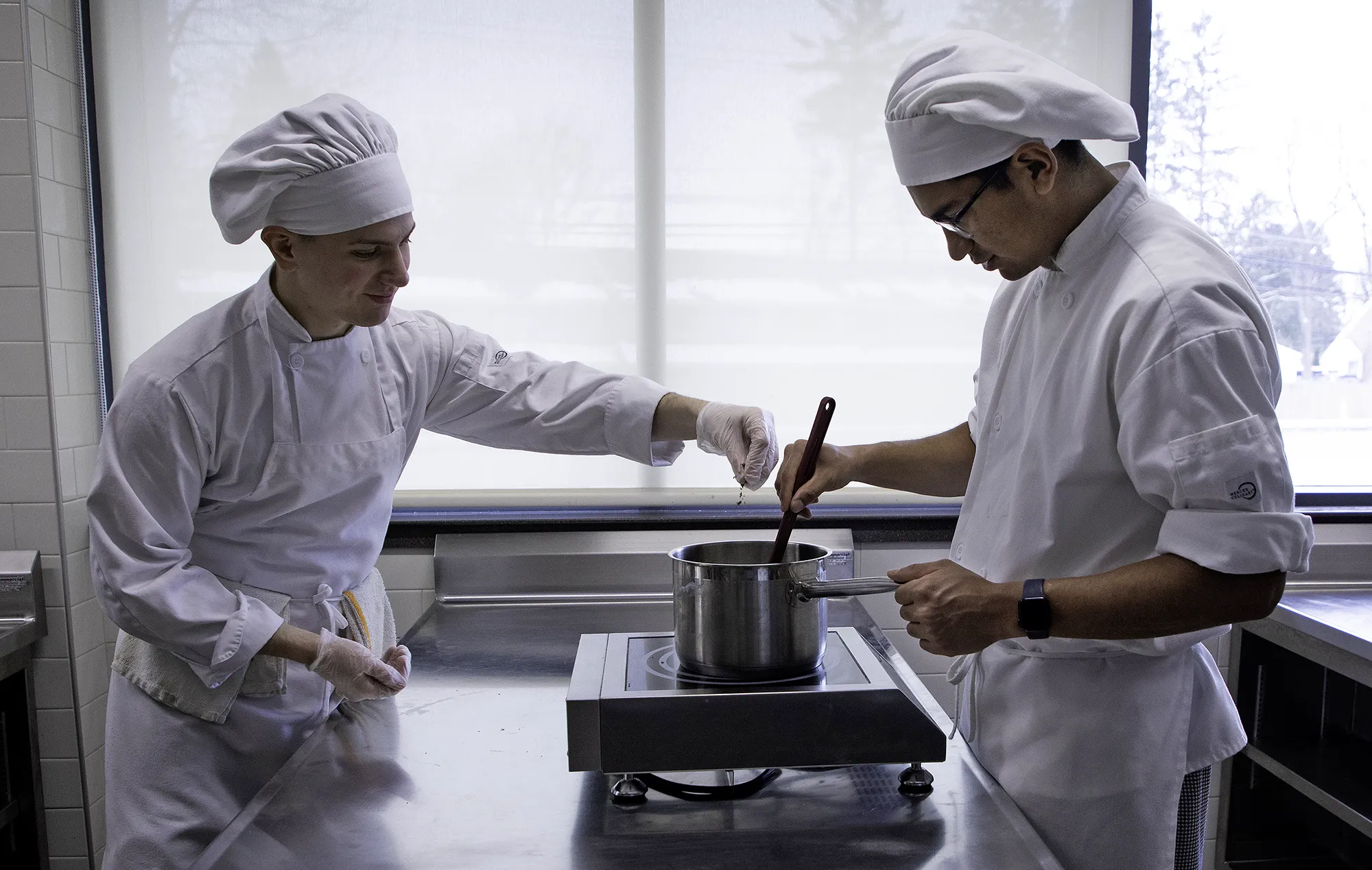 Two culinary arts students wearing white chef coats and hats work together at a stainless-steel workstation, stirring a pot on a countertop burner in a commercial teaching kitchen