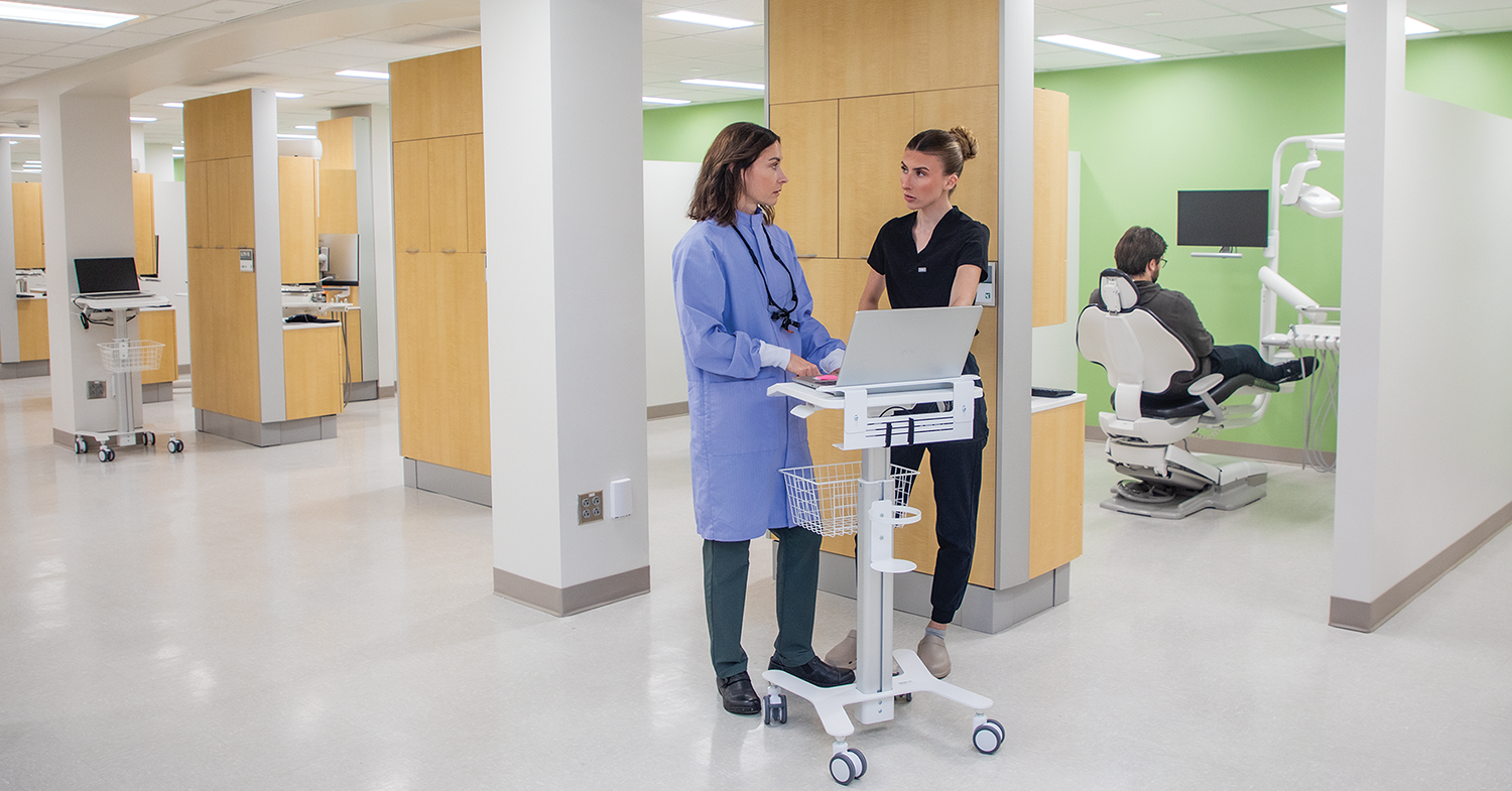 Dental Hygiene Two people standing by a workstation in a dental clinic with a patient sitting in a chair in the room behind them